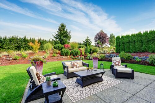 Die Gartenmöbel stehen auf einer Terasse aus Betonplatten. Sie ist von Rasen umgeben und bietet einen schönen Blick auf eine Blumen- und Baumgruppe.